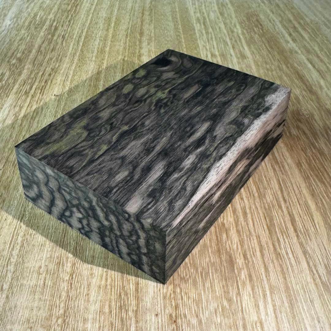 A rectangular pen-blank wood block showing dark and green peacock-burl grain placed on a light wooden surface.