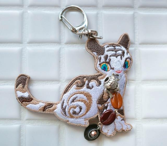 Embroidered cat keychain with decorative beads photographed on a white tiled background.