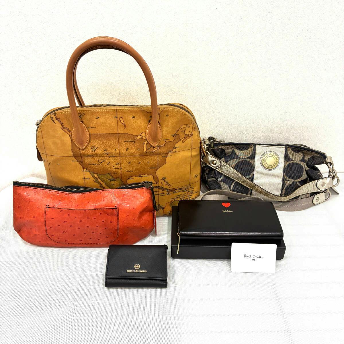 Five branded bags and wallets arranged on a white surface, including a Prima Classe map-print handbag, a Coach shoulder bag, an orange pouch, a Michael Kors small wallet, and a Paul Smith wallet with box.