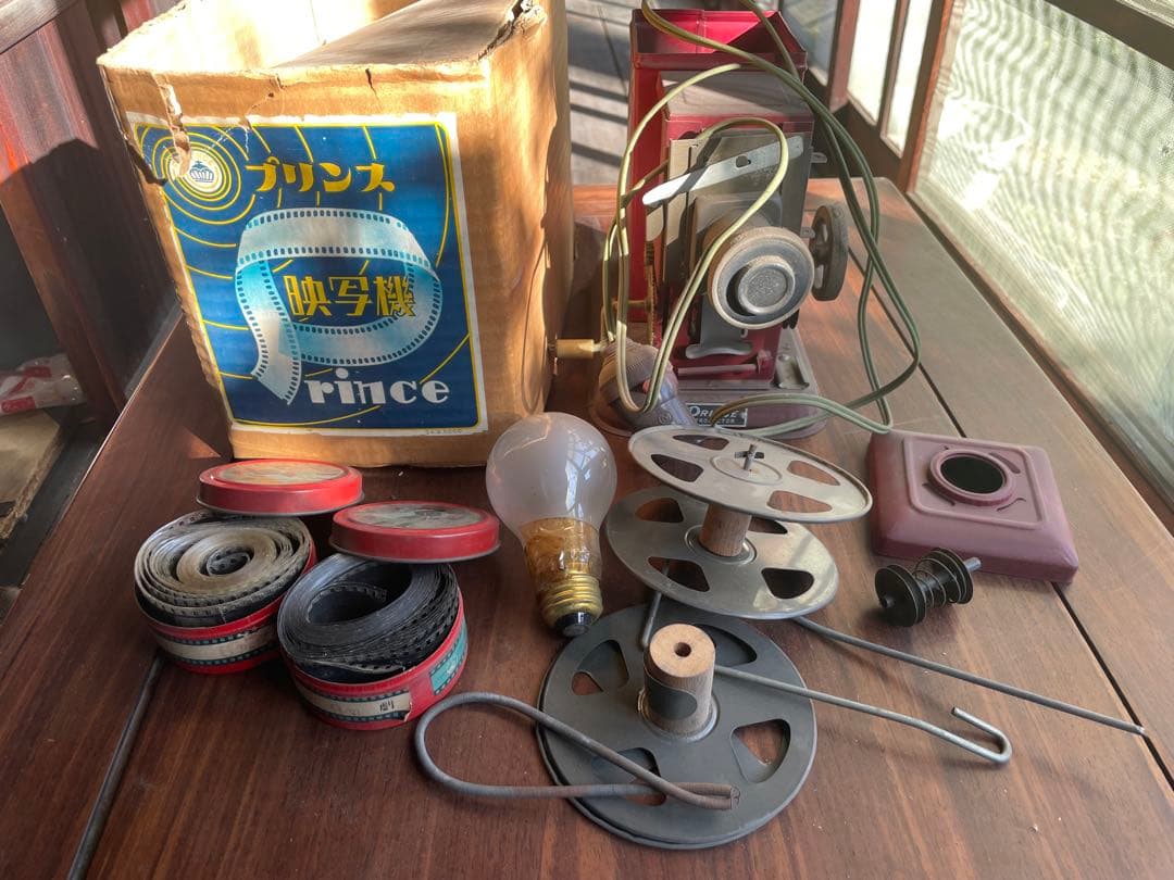 A Prince projector with film reels, film strips, a light bulb, and its box arranged on a wooden surface.