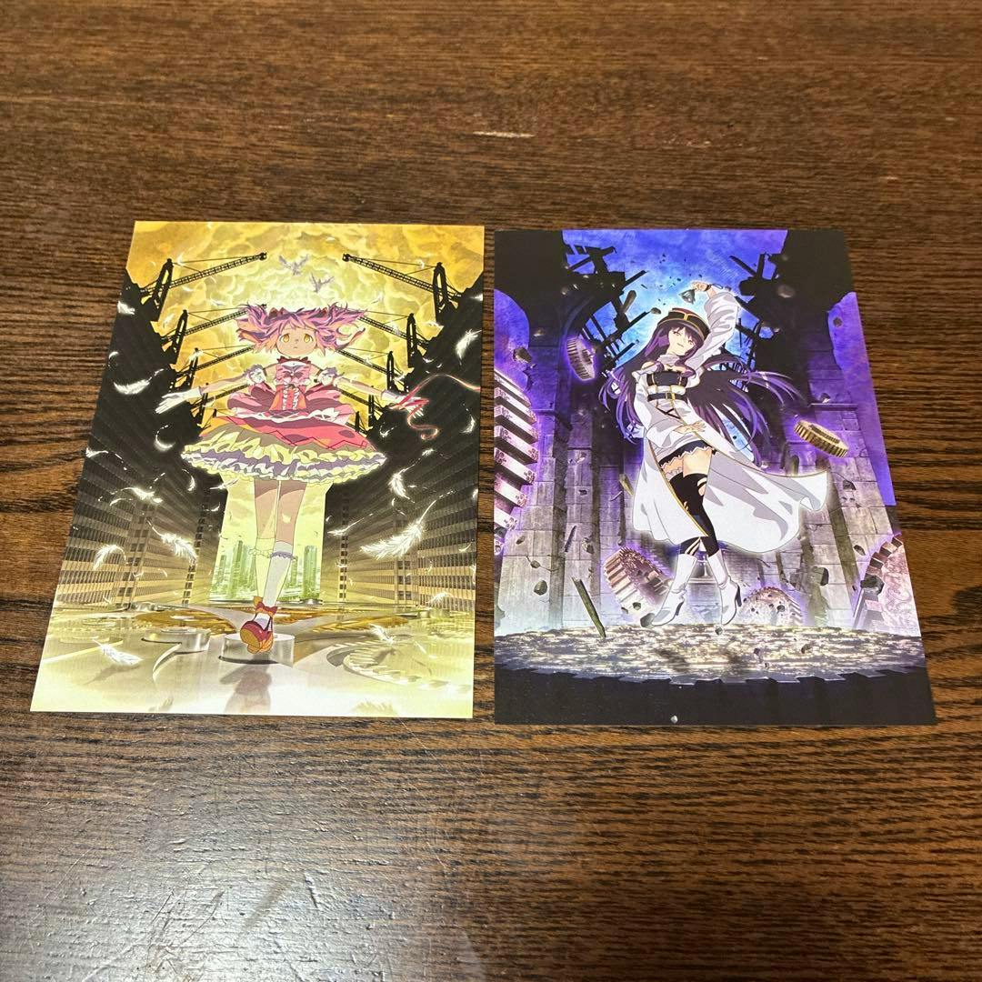 Two bonus postcards for 劇場版 魔法少女まどか☆マギカ ワルプルギスの廻天 shown side by side on a wooden table.