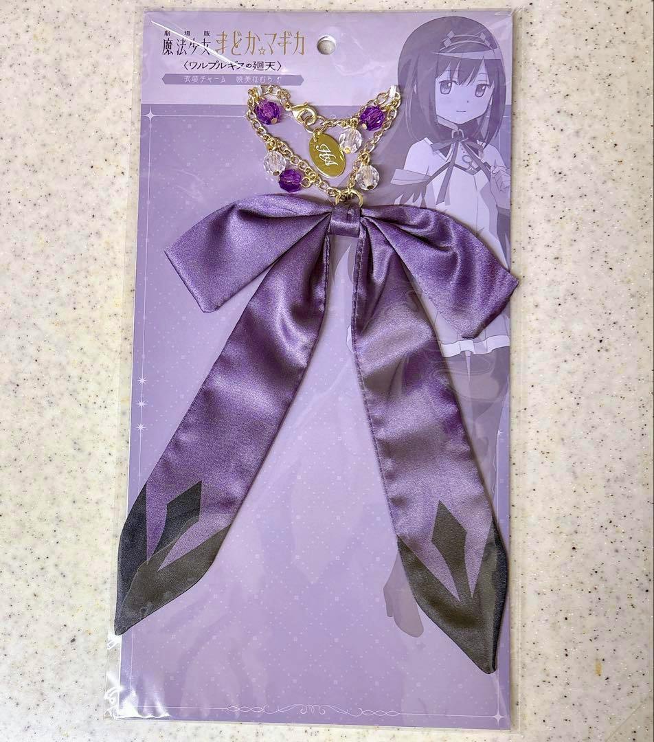 Purple fabric ribbon bag charm with gold chain and purple/clear beads on an illustrated Akemi Homura backing card, in sealed packaging.