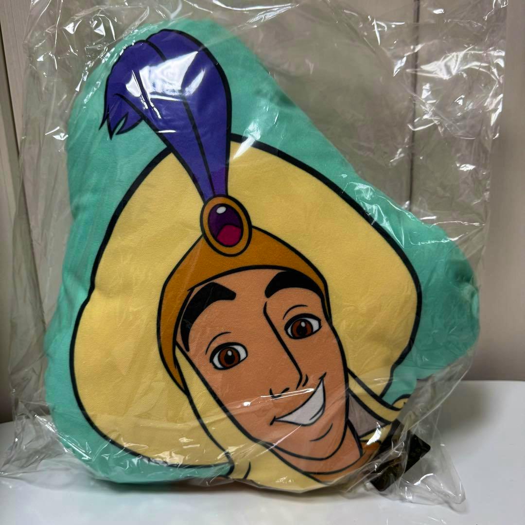 Front view of an Aladdin prince character plush cushion showing the character's face, sealed in plastic packaging.