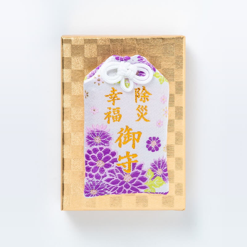A purple and white cloth omamori (amulet) with gold embroidered Japanese characters set on a gold presentation box.
