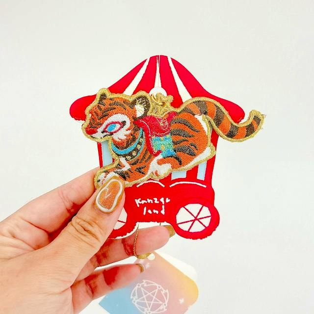 Hand holding an embroidered tiger patch charm keychain with a red carriage background.