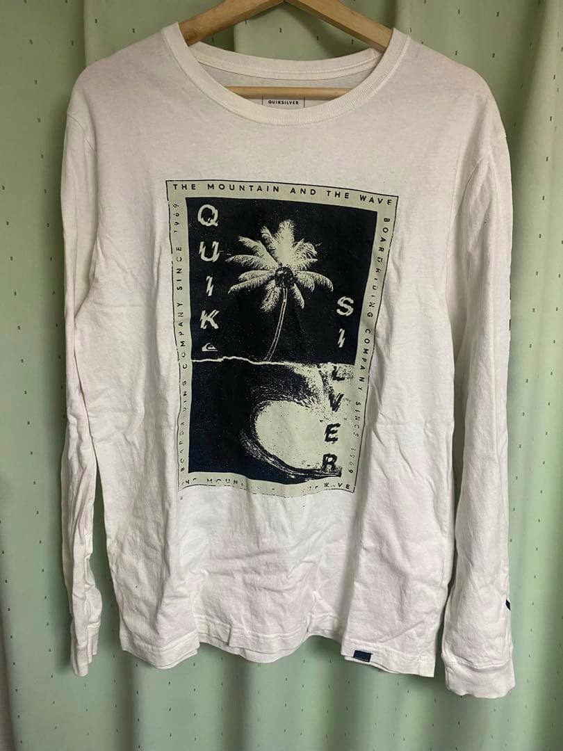 White men's long-sleeve QUIKSILVER cotton T-shirt hung on a wooden hanger showing a black palm tree and wave graphic on the front.