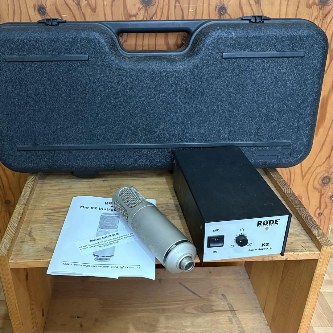 A RØDE K2 vacuum-tube condenser microphone shown with its power supply box, instruction manual, and hard carrying case on a wooden surface.