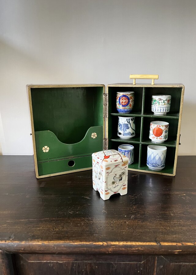 An open lacquered wooden box with green interior compartments holding small cups, with a small ceramic container placed in front.