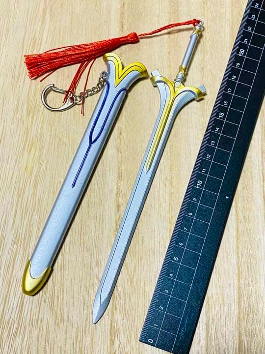 Photo of the Radiant Light (SAO) mini sword bag charm and its sheath with a red tassel and a ruler on a wooden surface.