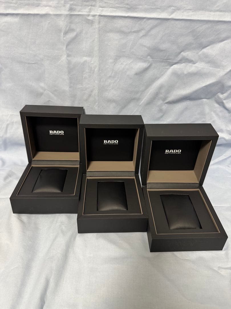 Three open RADO watch storage boxes arranged on a light cloth background.