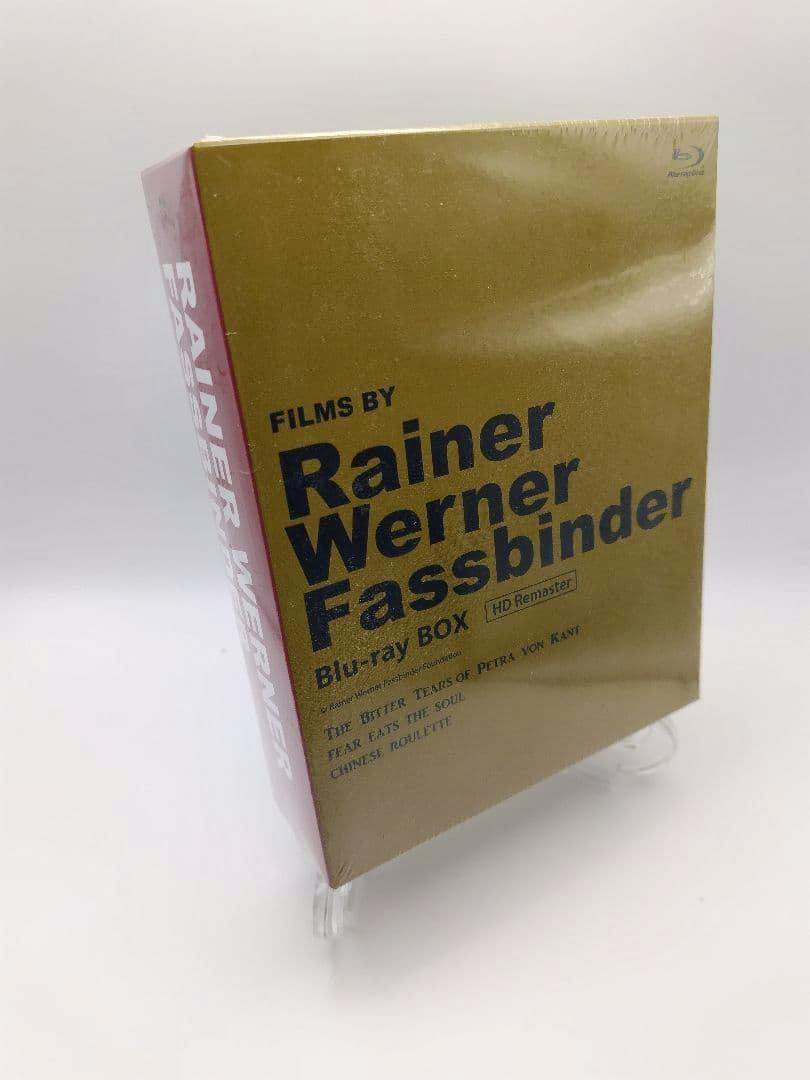 Shrink-wrapped gold-colored Rainer Werner Fassbinder Blu-ray box on a clear stand against a white background.
