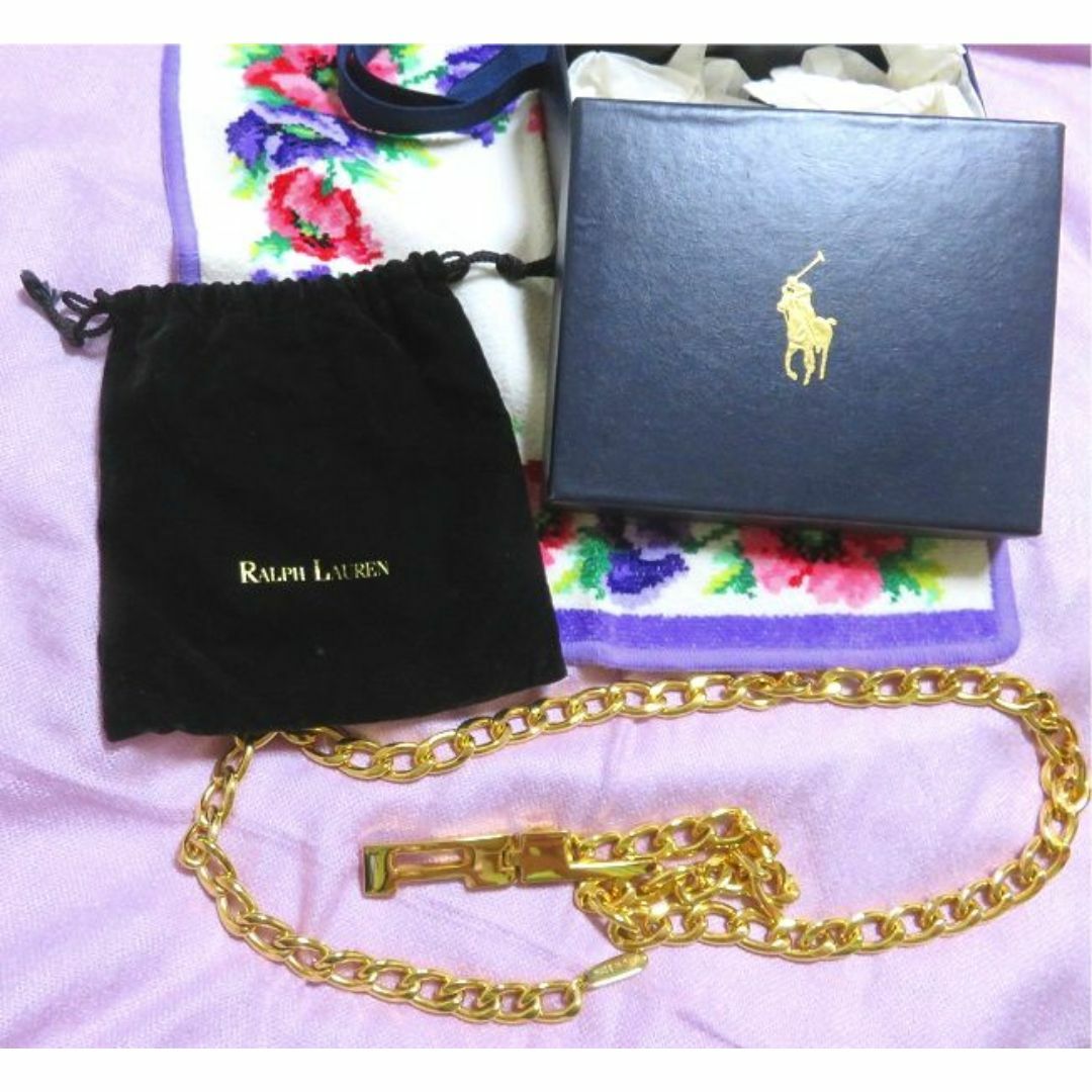 Gold chain belt displayed next to a Ralph Lauren box and black velvet pouch on a pink fabric background.