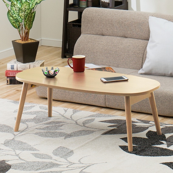 Oval wooden folding table in a natural finish placed on a living room rug in front of a sofa, with a red mug, small bowl, and smartphone on top.