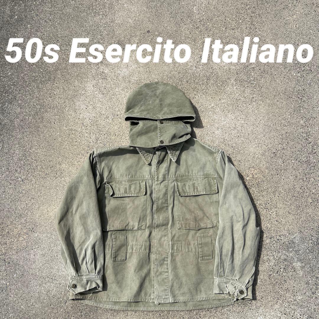 Short Italian military parachute jacket (men’s cotton woven) laid flat on concrete showing hood and front pockets, with white overlay text.