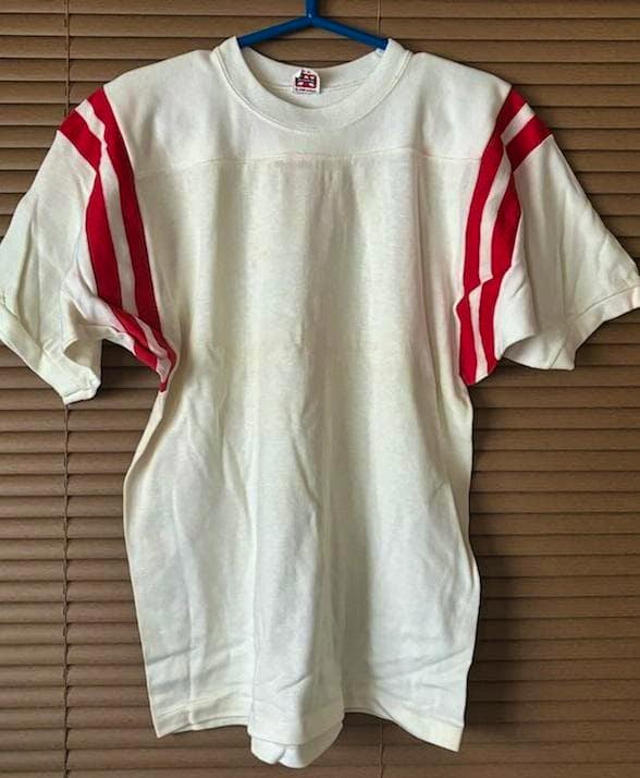 Front view of the vintage cotton football T-shirt with red stripes on the sleeves hanging on a blue hanger against a blind background.
