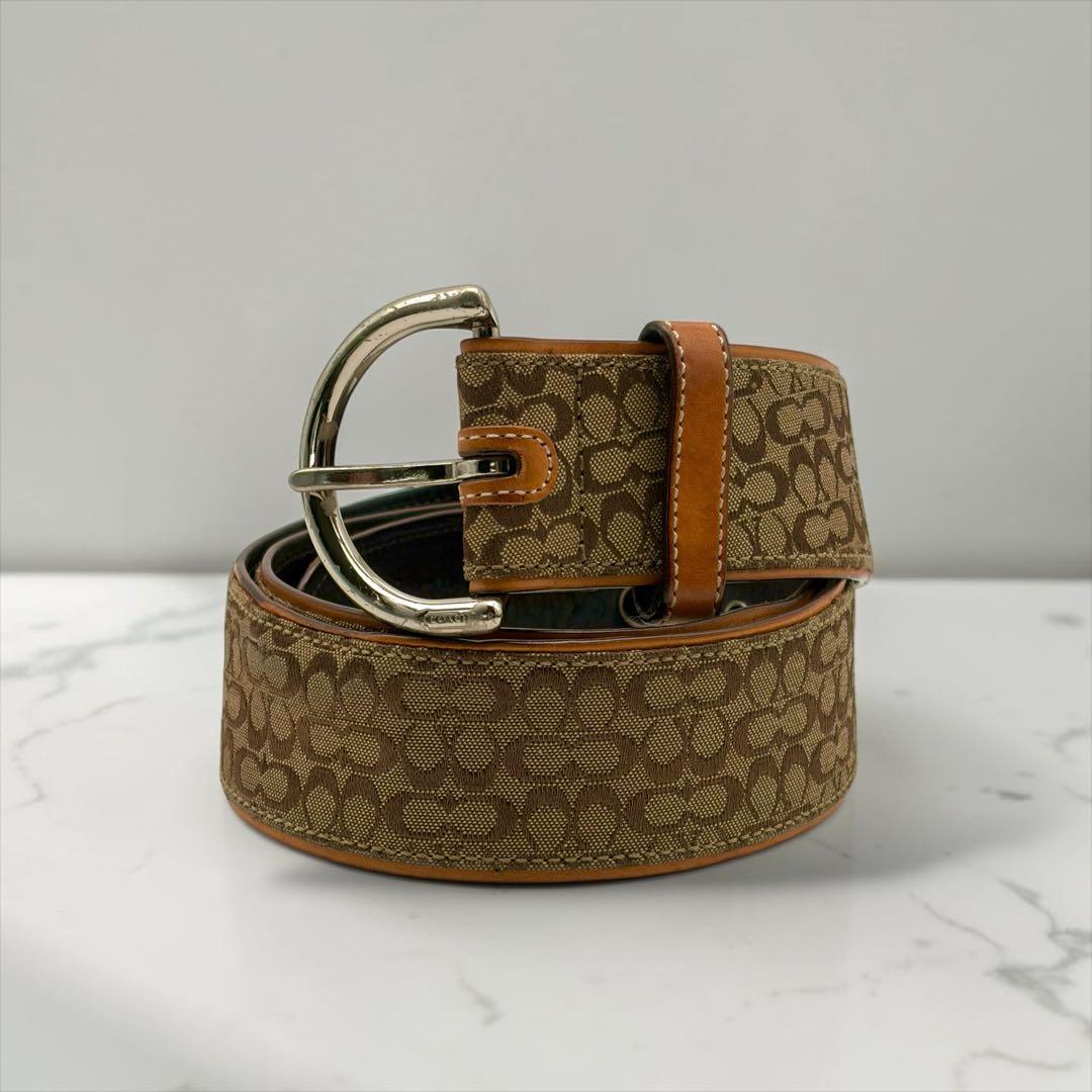 A rolled vintage Coach canvas belt with signature monogram pattern, leather trim, and silver buckle on a white surface.