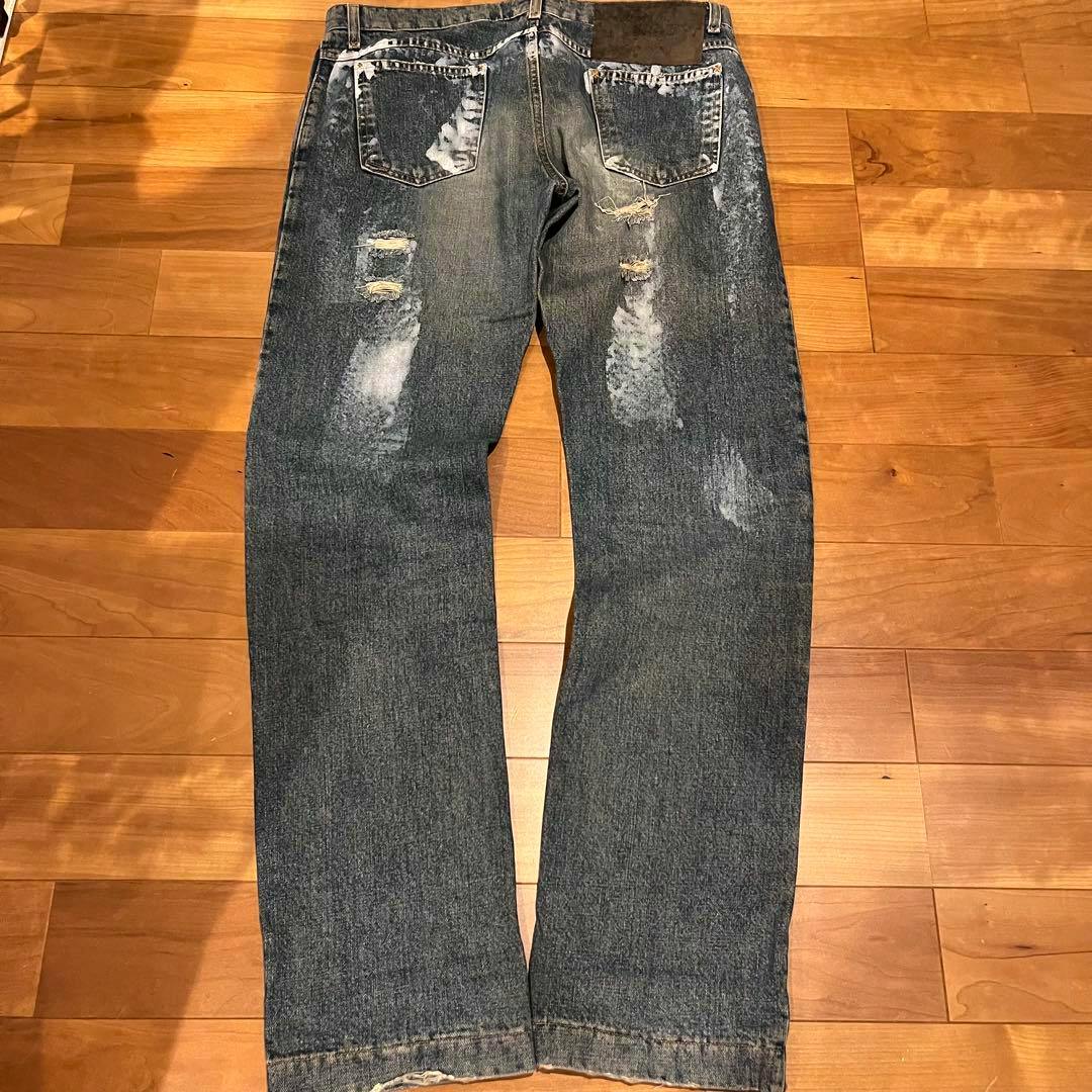 Back view of the D&G distressed paint-effect denim pants laid flat showing back pockets, rips, and paint marks on a wooden floor.