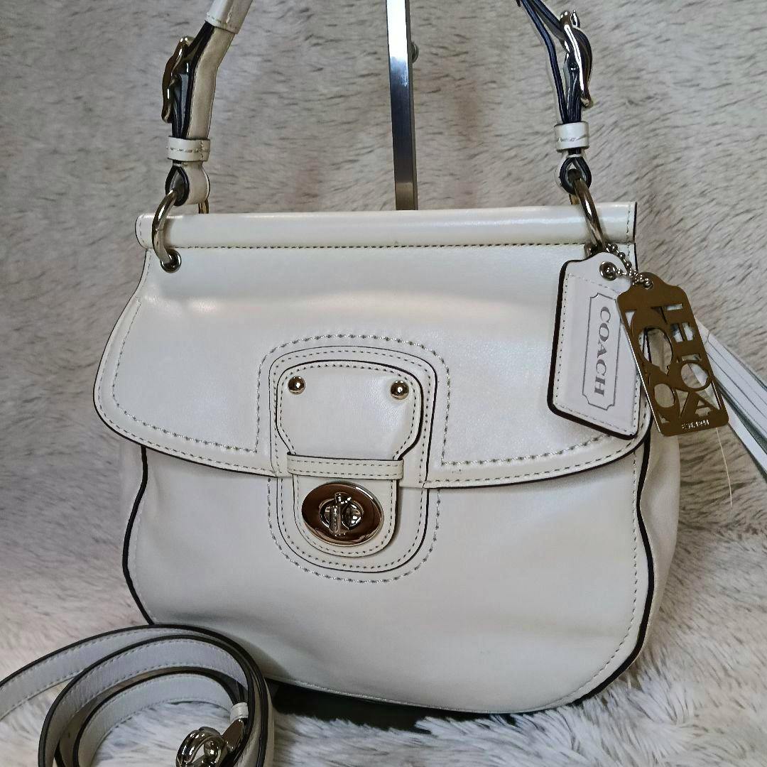 Front view of a white leather Coach handbag with a turnlock closure and a coiled shoulder strap visible.