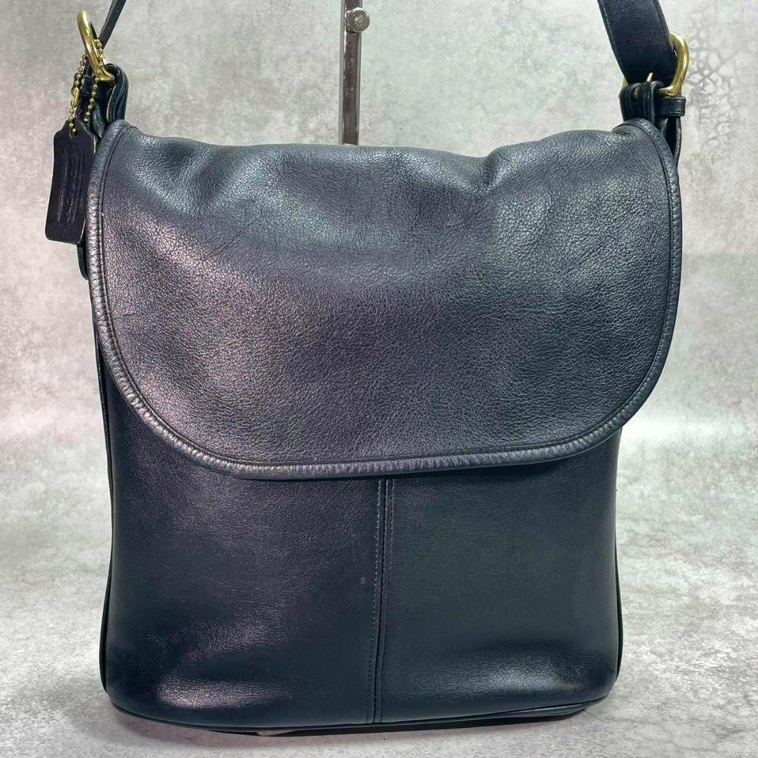 Front view of a black COACH leather shoulder bag (sacoche) with a flap closure, shoulder strap, and a leather hang tag.