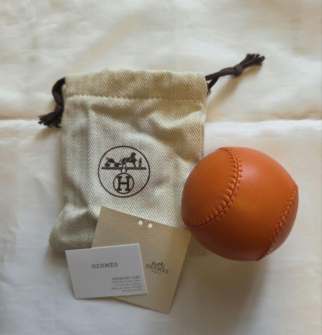 Leather orange baseball displayed next to a Hermes drawstring pouch and two cards on a light surface.