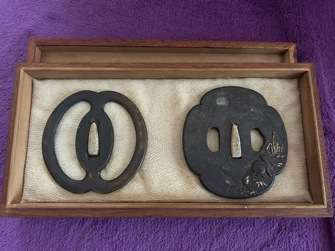 Two iron tsuba (Japanese sword guards) displayed side-by-side in a wooden paulownia box on a purple cloth.