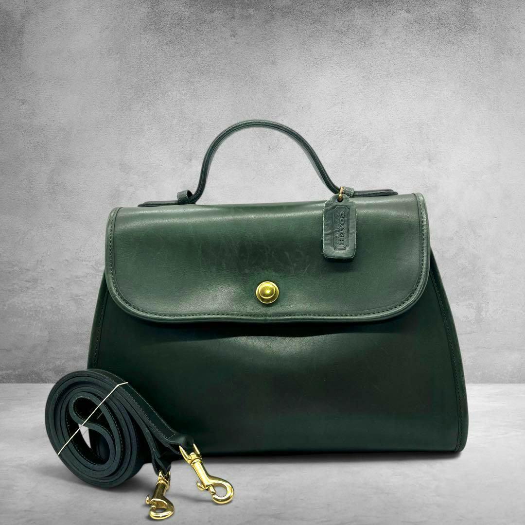 Dark green leather Coach 2-way handbag with top handle, front flap with gold-tone snap, and a coiled detachable shoulder strap placed in front.