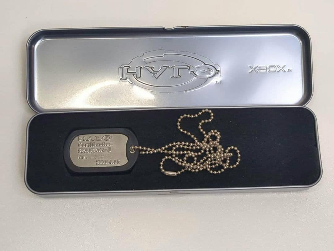 A Halo Spartan-II dog tag on a ball chain displayed inside an open embossed HALO Xbox metal tin case.