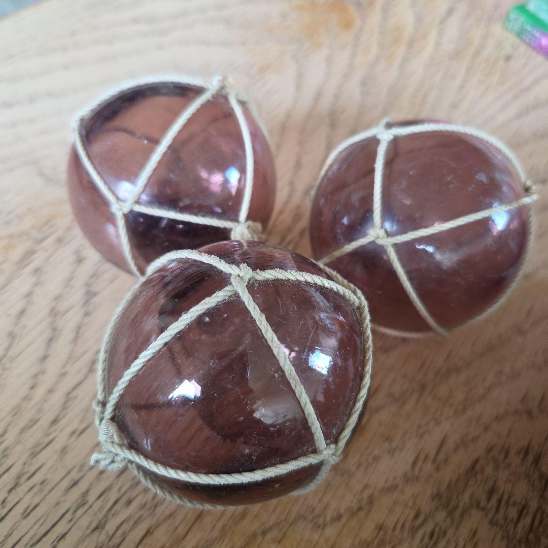 Three purple glass floats with knotted netting shown resting on a wooden surface.