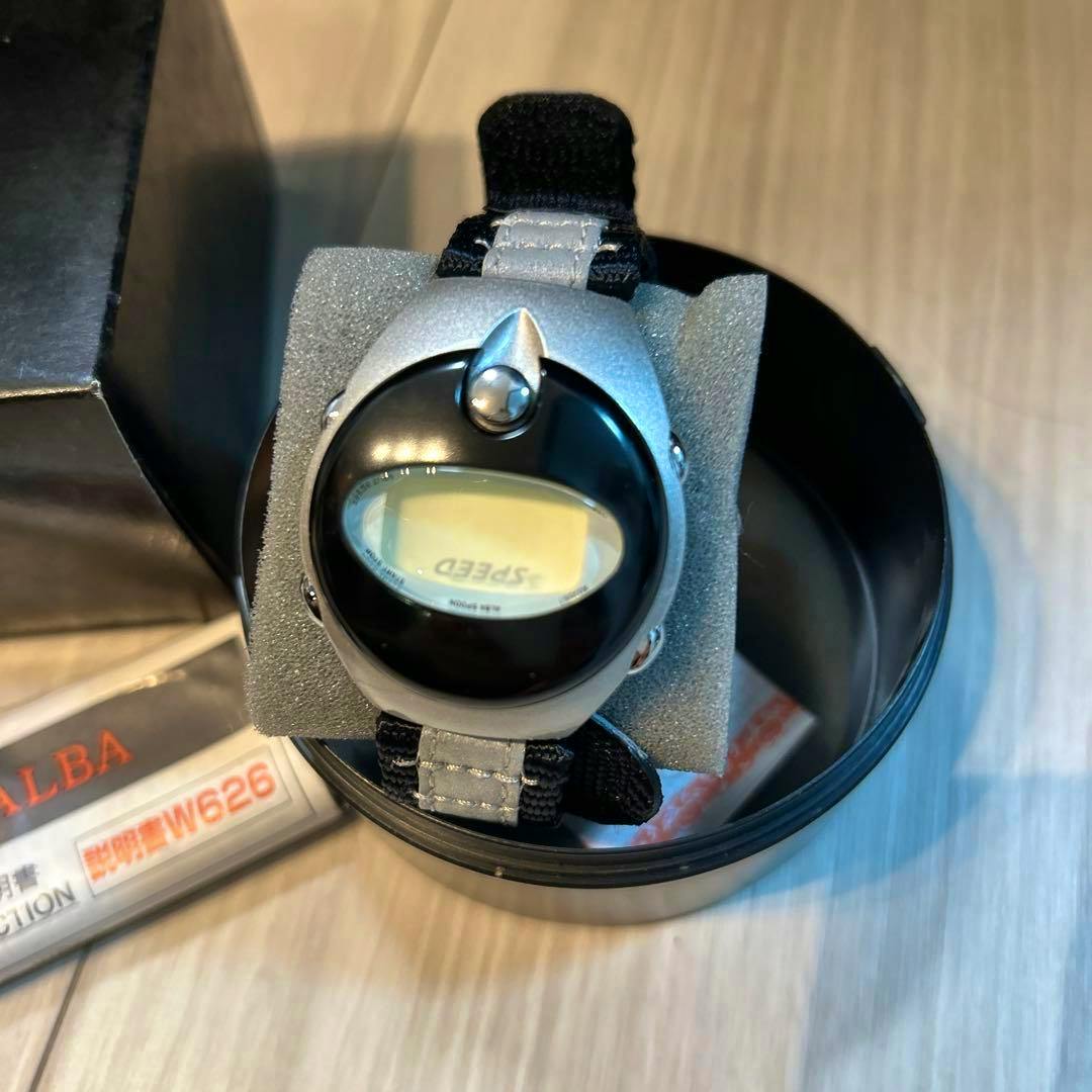 Close-up of the Seiko ALBA UFO SPEED digital wristwatch with a black bezel and strap displayed on foam inside its round tin.