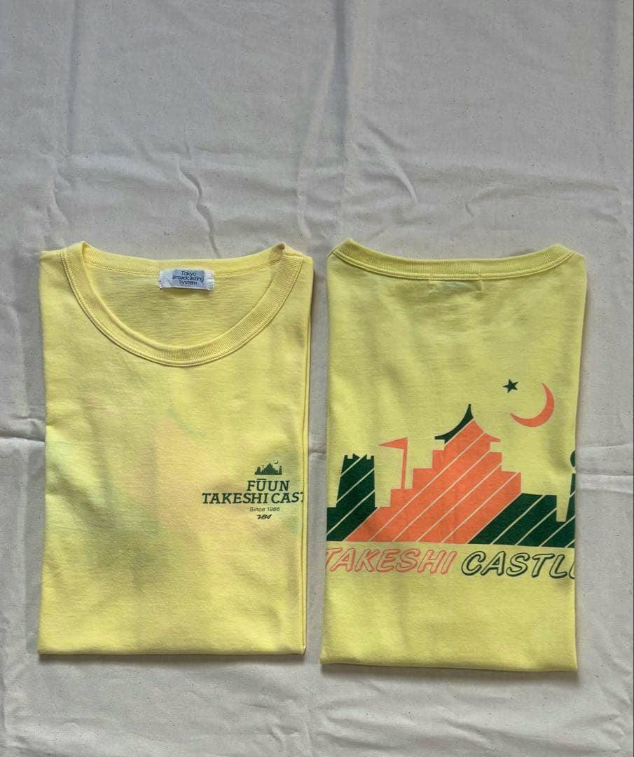 Two folded yellow Takeshi Castle T-shirts placed side by side, showing a small front logo on the left shirt and a larger castle graphic on the right.