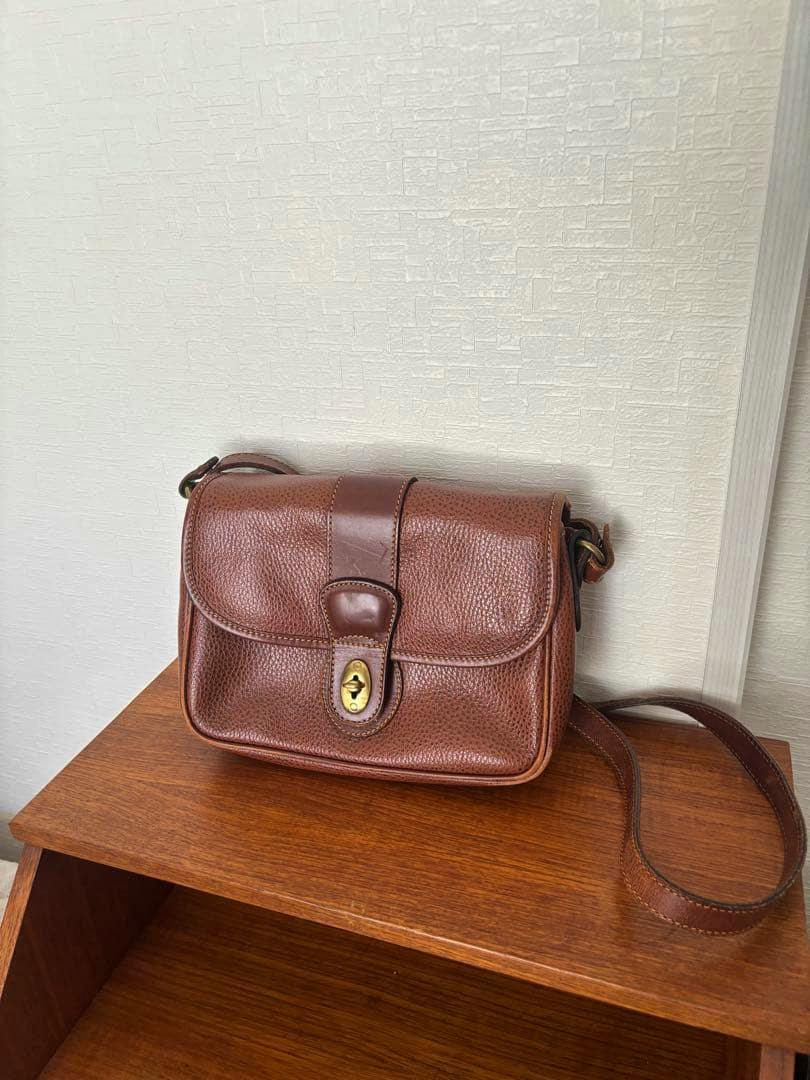 Brown Old Coach cow leather shoulder bag on a wooden surface, front view showing flap and turn-lock closure and shoulder strap.