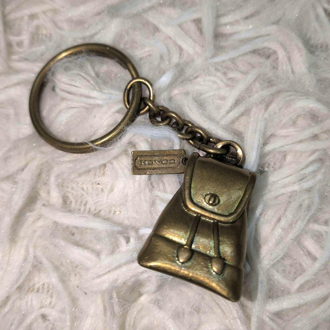 Coach backpack-shaped bag charm keyring in brass-toned zinc alloy on a white textured background.