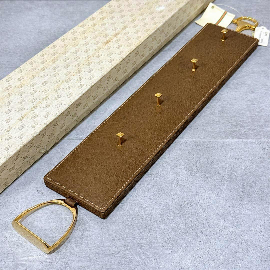 Brown leather belt hanger display with three gold-tone pegs and a gold stirrup-shaped hook shown alongside a patterned box.