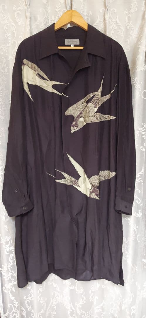 Long black silk Yohji Yamamoto pour homme shirt-dress with white swallow bird prints hanging on a wooden hanger against a curtain.