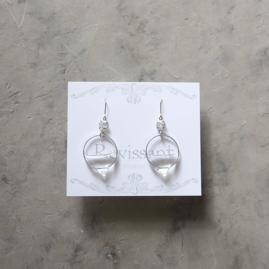 A pair of silver non-precious metal drop earrings with clear circular and teardrop elements displayed on a Ravissant card.