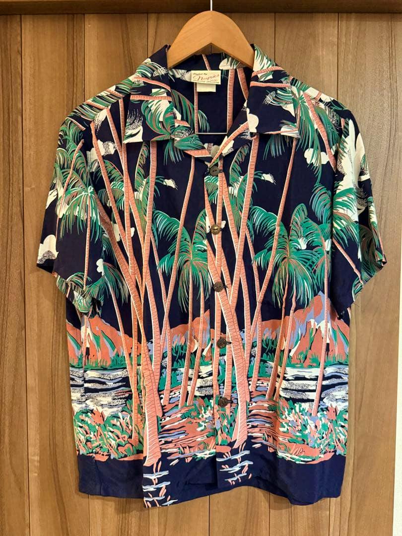 Front view of a men's rayon aloha shirt with a palm-tree pattern hanging on a wooden hanger against a wood-paneled background.