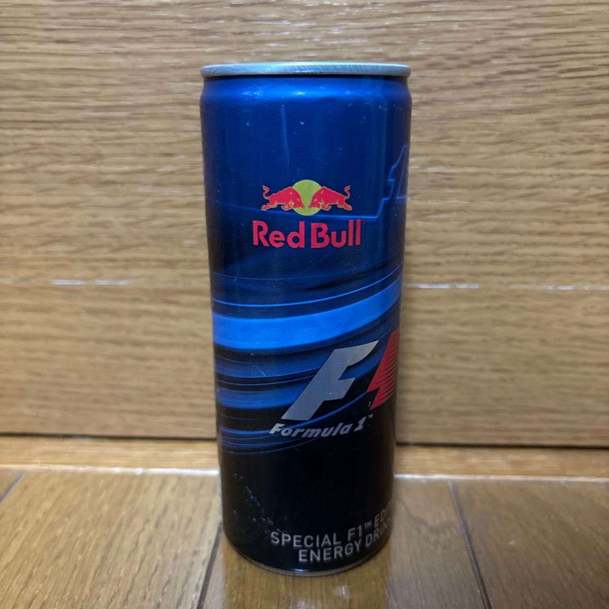 A Red Bull energy drink can with Formula 1 branding standing on a wooden floor against a wood backdrop.