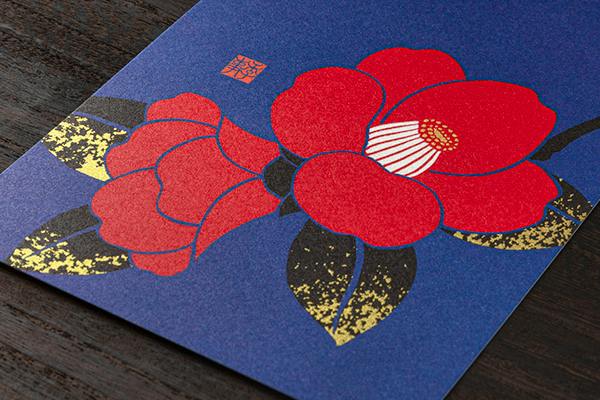 A postcard showing a red camellia floral design on a blue background placed on a wooden surface.