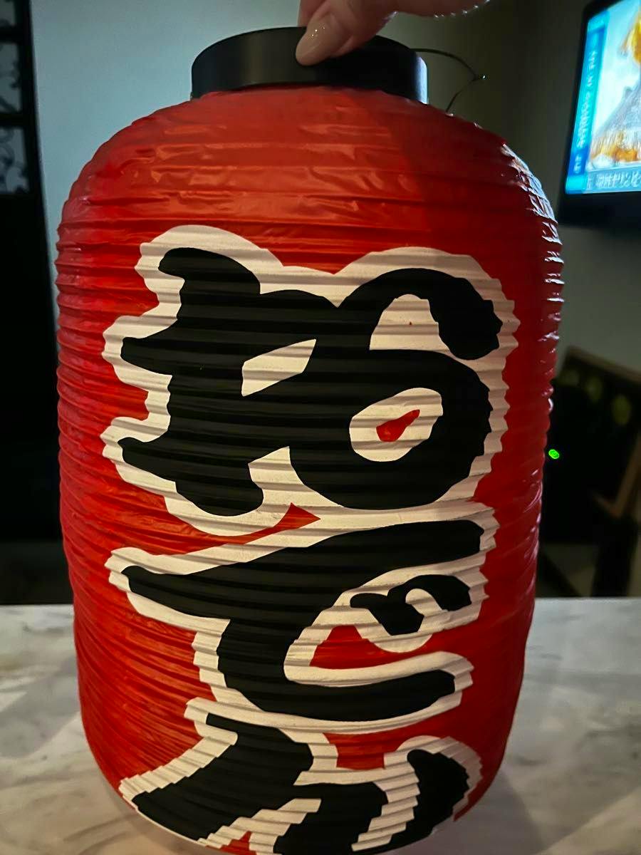 Red paper izakaya-style lantern with black-and-white kanji characters, shown upright and held at the top by a hand.