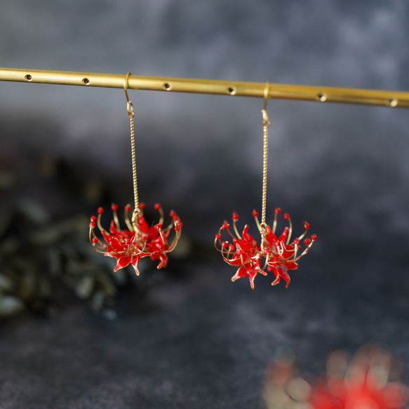 Pair of red spider-lily chandelier earrings hanging from a gold rod against a gray blurred background.