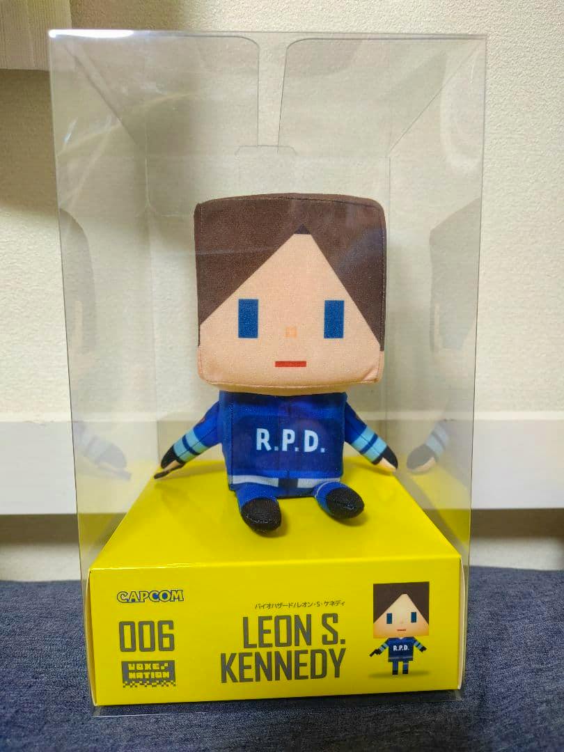 Front view of a boxed Voxenation Leon S. Kennedy plush in clear plastic packaging, showing the plush in an R.P.D. uniform and the yellow base labeled "LEON S. KENNEDY".