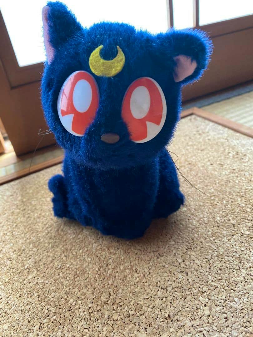 Front view of a retro Sailor Moon Luna plush toy with blue fur, red eyes, and a yellow crescent on its forehead sitting on a cork surface.
