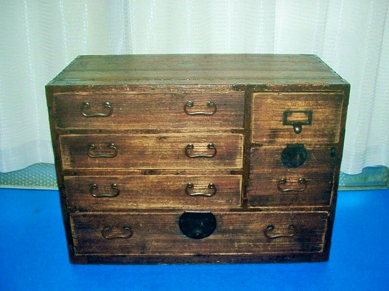 Front view of a retro small wooden chest (small tansu) with multiple drawers and copper metal handles placed on a blue surface.