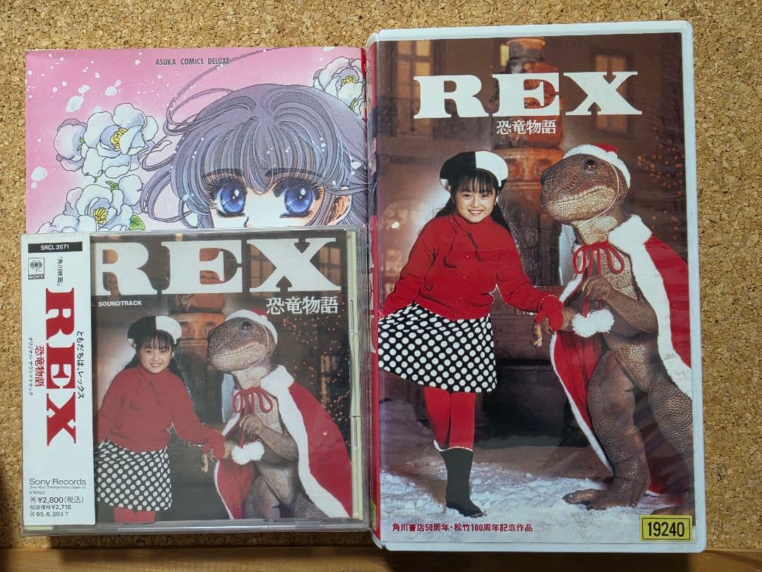 Front view of the REX 恐竜物語 VHS tape case displayed alongside the soundtrack and a comic on a cork surface.