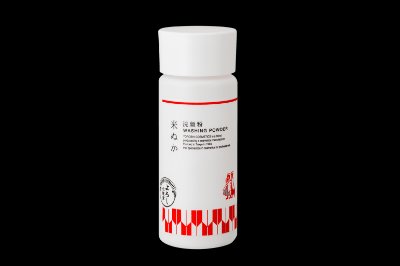 A white cylindrical container of 米ぬか洗顔粉 (rice bran face washing powder) with red decorative pattern shown against a black background.