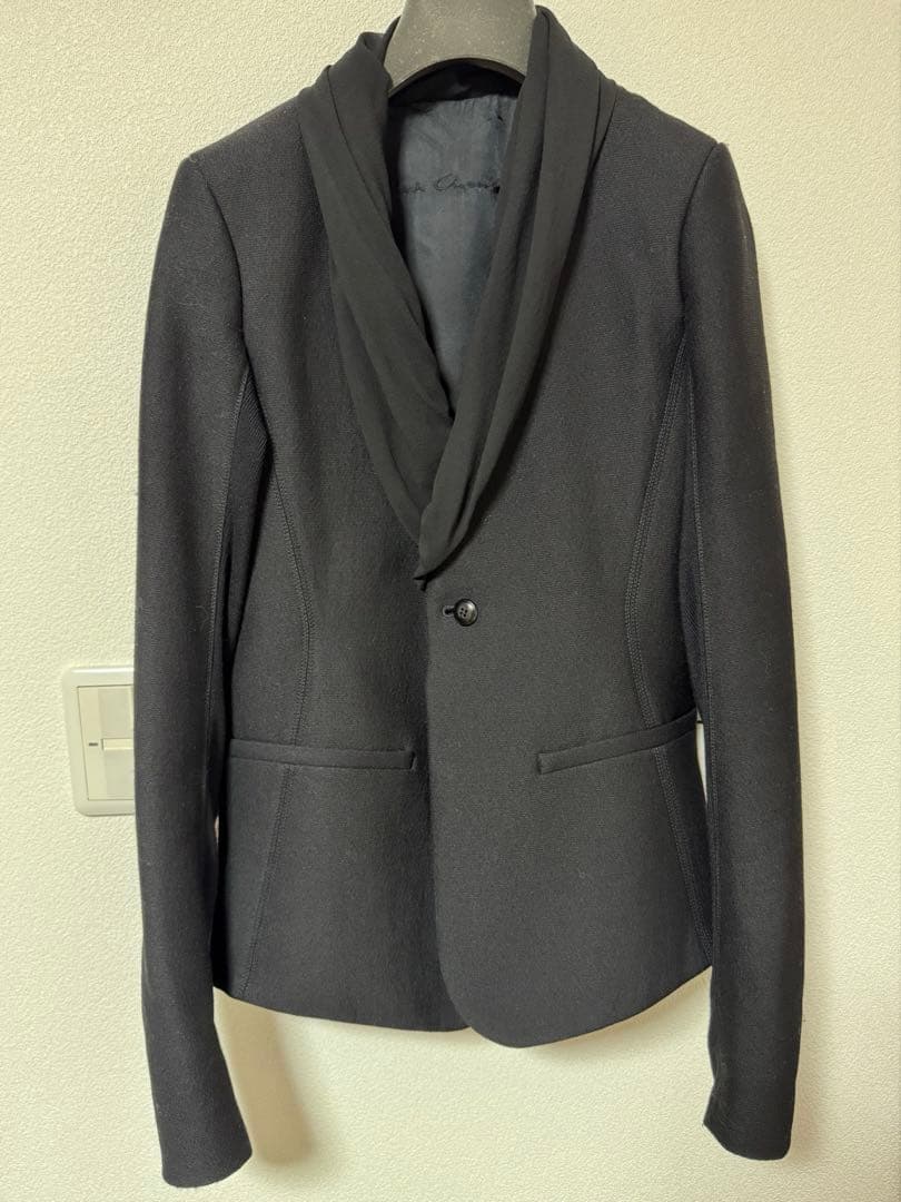 Front view of a black Rick Owens women's shawl-collar jacket hanging on a hanger against a light wall.