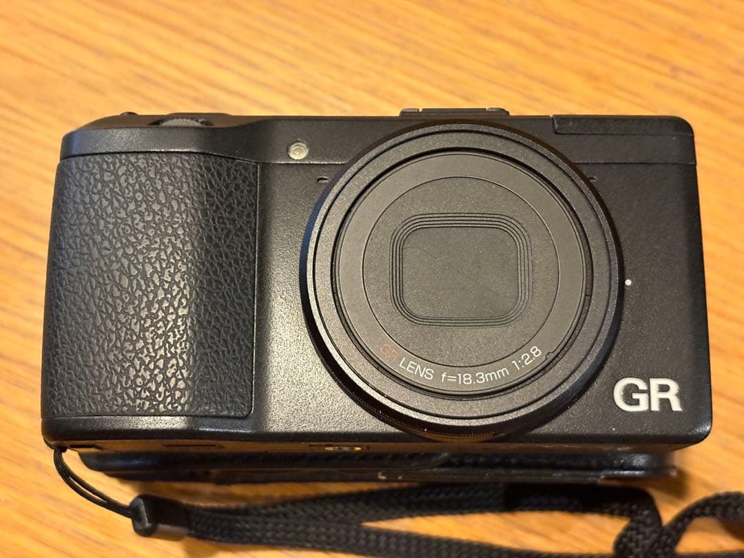 Front view of the RICOH GR APS-C digital camera with the lens closed and a wrist strap on a wooden surface.