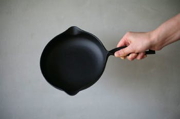 A black frying pan being held by a hand against a plain gray background.