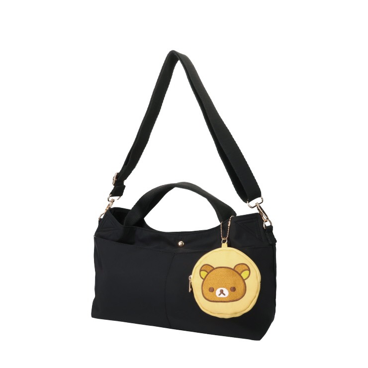 Black tote bag with short handles and a long adjustable shoulder strap, shown with a round Rilakkuma face coin pouch attached.