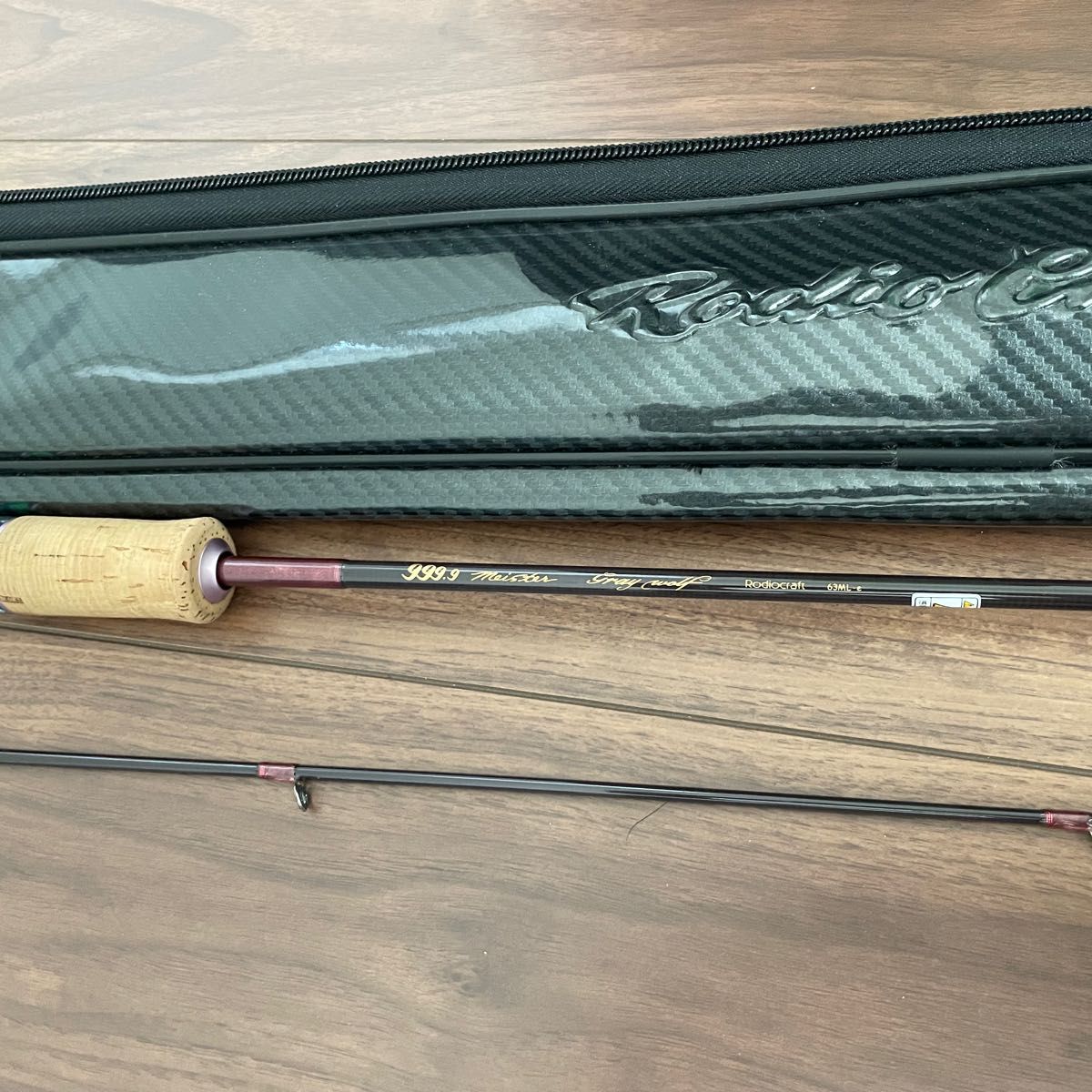 Sections of the ロデオクラフト 999.9 グレイウルフ63ML-e fishing rod and its carrying case laid on a wooden floor.
